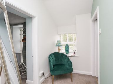 A green upholstered chair near a window with a lamp and vase on the sill and a mirror leaning against the wall at Bryn Peris in Moelfre