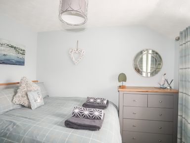 A bedroom with a bed, pillows, folded towels, a chest of drawers, a round mirror, and curtains at Bryn Peris in Moelfre