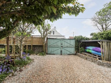 A gravel driveway with large closed blue wooden gates and a wooden shelter storing colorful kayaks at Bryn Peris in Moelfre