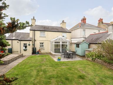A backyard with a grass lawn a patio table with chairs a small blue shed and a conservatory at Bryn Peris in Moelfre