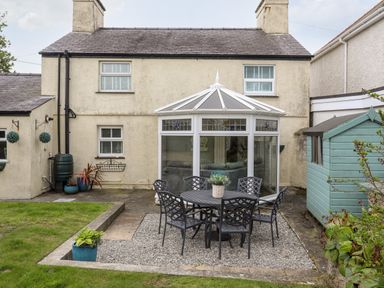 A backyard with a round table and six chairs on gravel in front of a conservatory and a blue shed at Bryn Peris in Moelfre