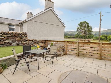 An outdoor patio area with a glass table and three chairs next to a wooden fence and stone wall with a house in the background at Bryn Hyfryd in Harlech