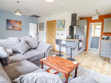 A living room with grey sofas a wooden coffee table and an open kitchen with bar stools at Bryn Hyfryd in Harlech