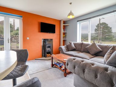 A living room with a grey sofa a wooden coffee table a wall-mounted TV and a stove at Bryn Hyfryd in Harlech