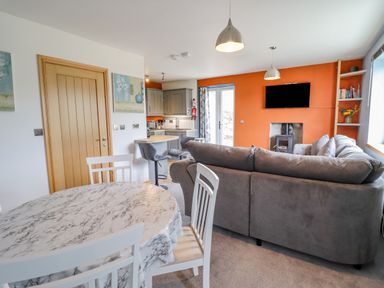 An open plan living room and kitchen with a round dining table and chairs a grey sofa orange accent wall and wall-mounted TV at Bryn Hyfryd in Harlech