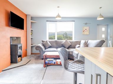 A living room with a grey sectional sofa a wood burning stove and a wall-mounted TV at Bryn Hyfryd in Harlech