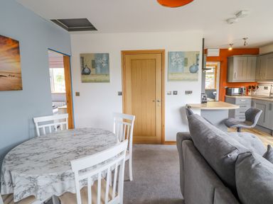 A dining area with a round table covered by a marble-pattern tablecloth and white chairs next to a living room with a gray sofa and an open kitchen with gray cabinets and an orange wall at Bryn Hyfryd in Harlech