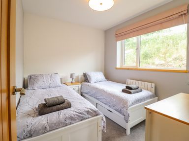 A bedroom with two single beds side by side with patterned bedspreads towels on each bed a small bedside table with a lamp a window with a blind and a chest of drawers at Bryn Hyfryd in Harlech