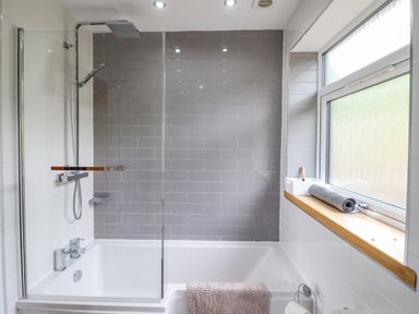 A bathroom with a bathtub shower combination grey tiled wall and a window sill with a rolled mat at Bryn Hyfryd in Harlech