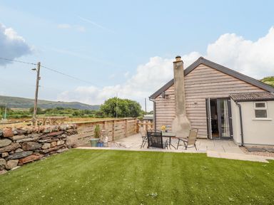 A backyard with a patio table and chairs next to a house with wooden siding and a stone chimney at Bryn Hyfryd in Harlech