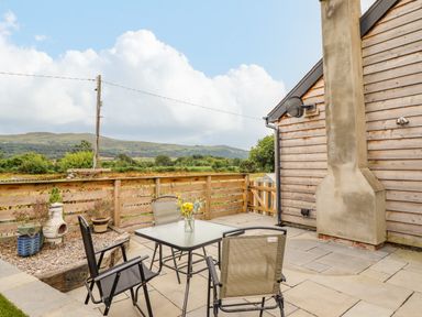 An outdoor patio with a glass table and four chairs next to a wooden fence and a building wall with a chimney at Bryn Hyfryd in Harlech