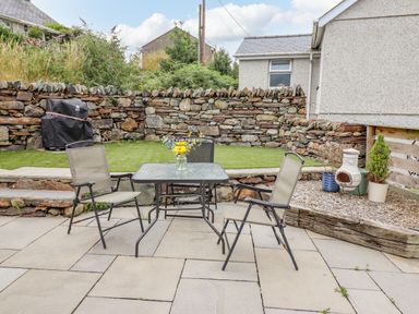 An outdoor patio with a glass table and three chairs a stone wall and a covered barbecue Bryn Hyfryd Harlech