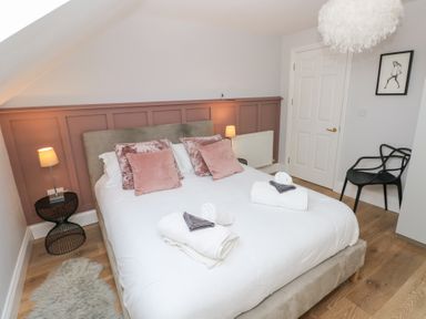A bedroom with a double bed white bedding pink cushions bedside tables lamps a black chair and a door at Cambrie in Tenby