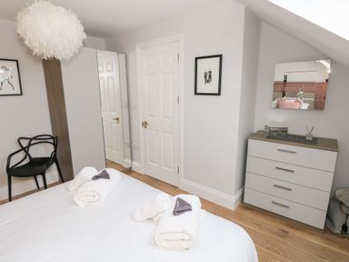 A bedroom with a bed, white towels, a black chair, a wardrobe with a mirrored door, a white door, framed pictures, a chest of drawers with a mirror, and wooden flooring at Cambrie in Tenby