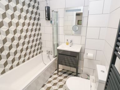 A bathroom with a bathtub shower combination a sink with a mirror above a toilet and geometric patterned tiles at Cambrie in Tenby