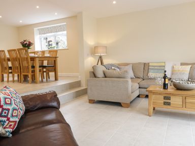 A living room with a beige sectional sofa a wooden coffee table with wine bottles glasses and a bowl a wooden dining table with chairs and flowers on it at Hen Dy Bryn Mawndy Melin y Wig near Corwen