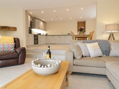 An open living area with a beige sofa and cushions a wooden coffee table with a bowl a brown leather armchair a kitchen with grey cabinets and a dining table with flowers at Hen Dy Bryn Mawndy Melin y Wig near Corwen