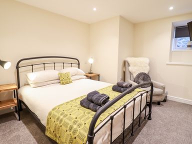 A bedroom with a metal bed frame a yellow patterned runner and folded towels on the bed a nightstand with a lamp an armchair with a cushion and throw and a small window at Hen Dy Bryn Mawndy Melin y Wig near Corwen