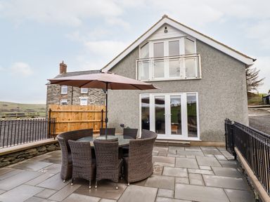 A patio with a round table and wicker chairs under an umbrella outside a two-story house with a balcony at Hen Dy Bryn Mawndy Melin y Wig near Corwen