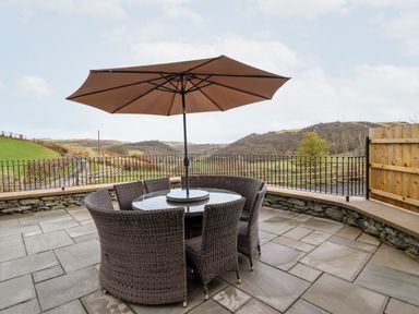 An outdoor patio with wicker dining chairs a glass table and a large umbrella overlooking a rural landscape at Hen Dy Bryn Mawndy Melin y Wig near Corwen