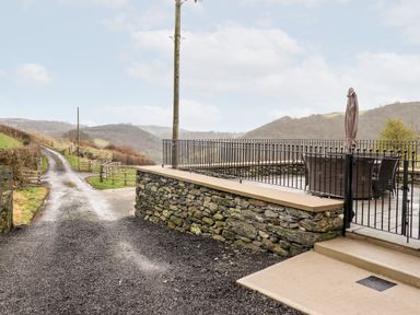 An outdoor patio area with a table and umbrella behind a stone wall and metal fence next to a gravel road with hills in the background at Hen Dy Bryn Mawndy Melin y Wig near Corwen