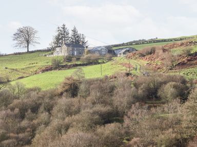 A rural farmhouse with surrounding outbuildings on a grassy hill above a wooded area at Hen Dy Bryn Mawndy in Melin y Wig near Corwen