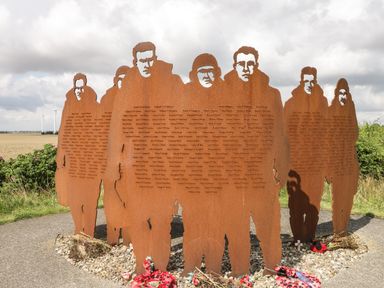 Rust-colored metal sculptures of soldiers with names inscribed on them standing on a pebble base outdoors with fields and wind turbines in the background