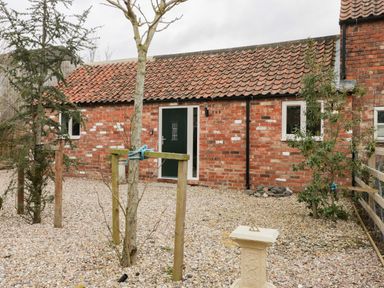 A gravel courtyard with small trees and a sundial in front of a brick building with a tiled roof and green door at Robins Nest Cottage in Brandesburton