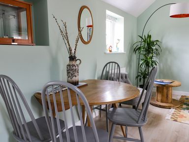 A dining area with a round wooden table and four gray chairs a vase with branches a window a round wooden side table with books a floor lamp and a plant at Robins Nest Cottage Brandesburton