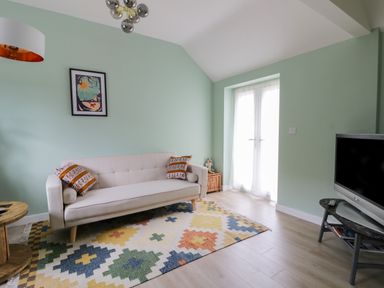 A living room with a beige sofa with patterned pillows a colorful geometric rug a TV on a stand and a round wooden side table at Robins Nest Cottage in Brandesburton