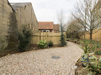 A gravel path bordered by plants and trees with a wooden fence and buildings in the background at Robin's Nest Cottage in Brandesburton
