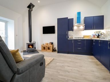 A living area with a dark blue kitchen, wood-burning stove, TV on the wall, and gray armchair at 3 Mountain View in Talwrn near Llangefni