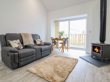 A living room with a grey sofa, a small wooden dining table with two chairs, a beige rug, and a wood burning stove at 3 Mountain View in Talwrn near Llangefni