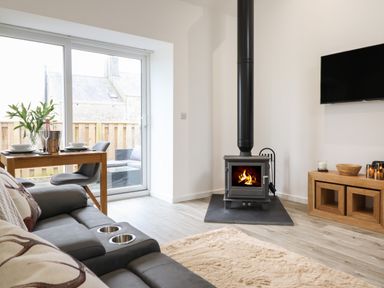 A living room with a wood stove a sofa a dining table with chairs and a wall-mounted television at 3 Mountain View in Talwrn near Llangefni