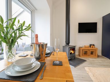 A dining table set with plates glasses and a champagne bottle in a bucket near a wood stove and a wall mounted television at 3 Mountain View Talwrn near Llangefni