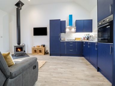 A kitchen with blue cabinets a stove and oven next to a wood-burning stove and a wall-mounted TV at 3 Mountain View in Talwrn near Llangefni