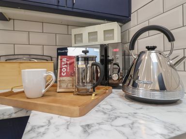 A kitchen counter with a wooden tray holding a white mug a bag of costa coffee and a french press next to a microwave and a stainless steel kettle at 3 Mountain View in Talwrn near Llangefni
