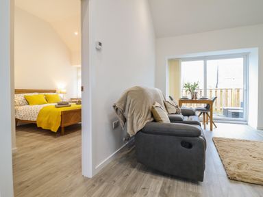 A living area with a gray sofa and a wooden dining table with chairs next to a sliding glass door and a bedroom with a bed and yellow bedding at 3 Mountain View in Talwrn near Llangefni