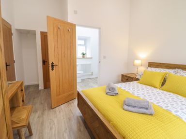 A bedroom with a wooden bed with yellow bedding two wooden nightstands a lamp and an open door leading to a bathroom at 3 Mountain View in Talwrn near Llangefni