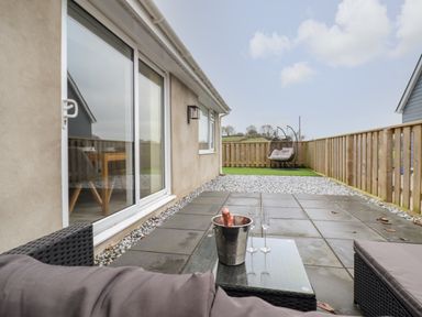 A fenced patio area with outdoor seating a glass table with a bucket and two glasses and a hanging chair on artificial grass at 3 Mountain View in Talwrn near Llangefni