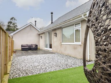 An outdoor patio area with a gray cushioned sofa on a tiled section and a hanging wicker chair on a grassy area at 3 Mountain View in Talwrn near Llangefni