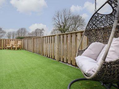 An outdoor area with artificial grass a hanging chair with cushions and two wooden chairs behind a wooden fence at 3 Mountain View in Talwrn near Llangefni