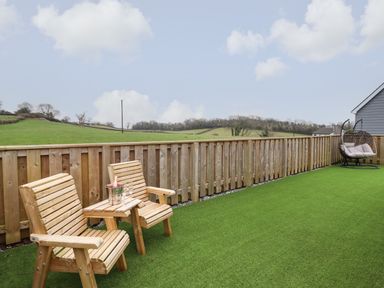 An outdoor patio area with two wooden chairs and a table with a bottle in an ice bucket and two glasses and a hanging chair at 3 Mountain View in Talwrn near Llangefni