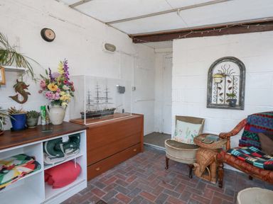 A room with brick flooring, a wooden cabinet with a model ship, a white shelf with blankets and plants, chairs with cushions and blankets, and wall decorations at Victorian Town House in Weymouth