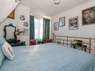 A bedroom with a brass bed frame a blue patterned bedspread framed floral pictures on the wall a black dresser with a mirror and two pink chairs by the window at Victorian Town House in Weymouth