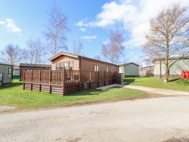 An exterior view of a wooden lodge with a deck surrounded by grass and trees at South Lakeland Leisure Village