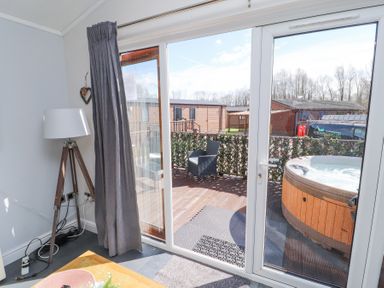 A room with a floor lamp and a wooden table next to glass sliding doors leading to a deck with a chair and a hot tub at Duckling Lodge in South Lakeland Leisure Village
