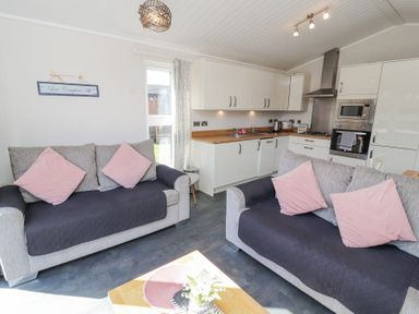 A small living area with two grey sofas with pink cushions next to a kitchen with white cabinets and wooden countertops at Duckling Lodge in South Lakeland Leisure Village