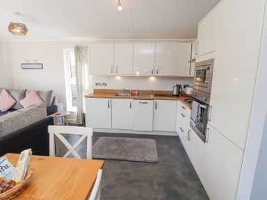 A kitchen with white cabinets wooden countertop and gray rug next to a living room with gray sofa and pink cushions at Duckling Lodge in South Lakeland Leisure Village