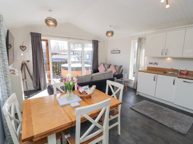 A dining table with white chairs and flowers near a living room and kitchen at Duckling Lodge in South Lakeland Leisure Village
